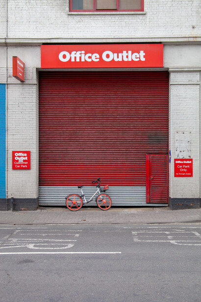 Closed storefront with red corrugated metal security shutter and Office Outlet signage, bicycle parked in front.