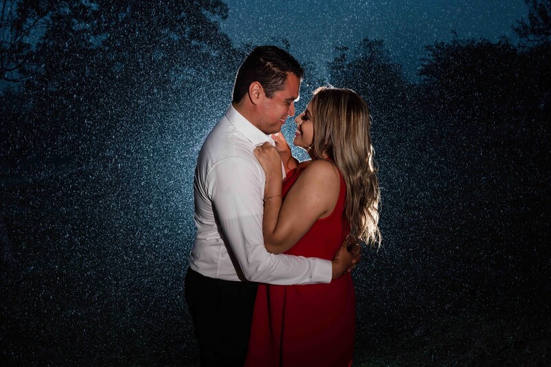 Couple embracing in the rain against a dark background with trees.
