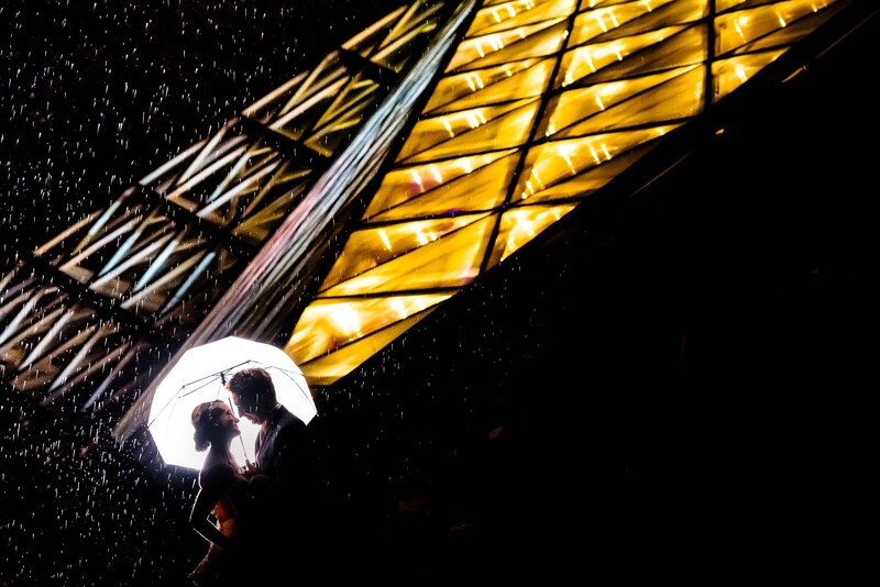 Silhouetted couple holding umbrella in heavy rain beneath warm golden architectural lighting at night.