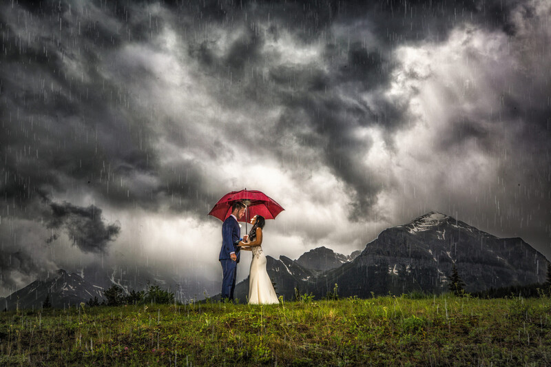 Bride and groom standing under a red umbrella in a storm with dramatic dark clouds overhead.
