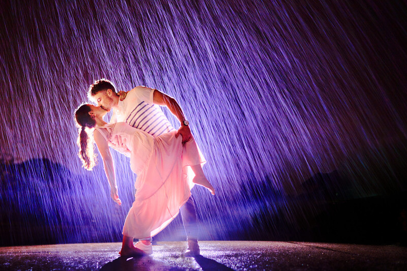 Man lifting woman in flowing pink dress under dramatic purple rain effect lighting.