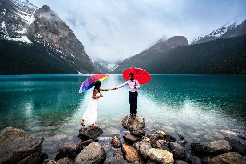 Couple holding colorful umbrellas on rocks at the edge of a turquoise alpine lake surrounded by snow-capped mountains.