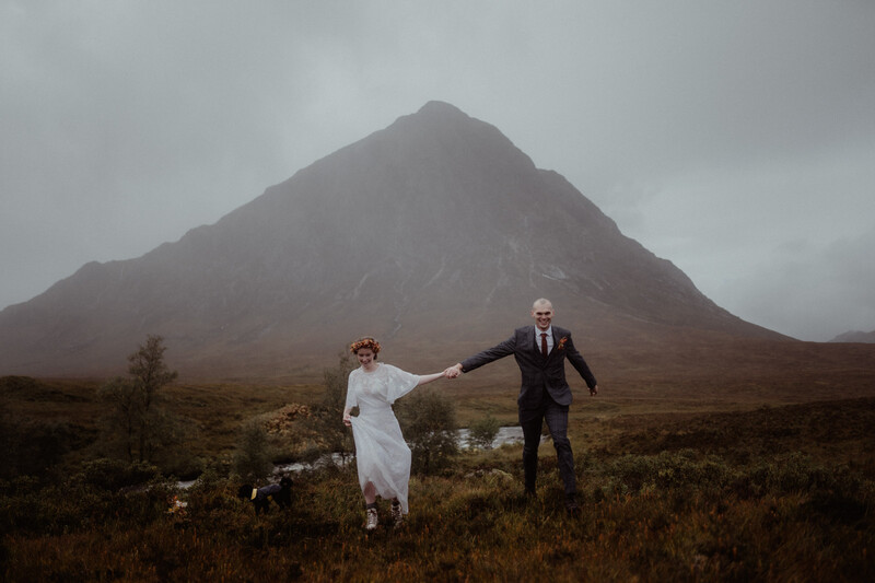 Bride and groom holding hands in a misty moorland valley with a dramatic peaked mountain behind them.