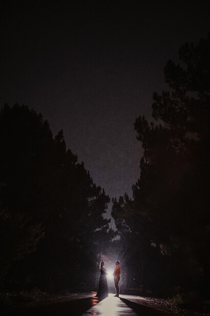 Two figures standing on a road at night, flanked by dark silhouettes of tall trees, with a single light source creating a dramatic glow on the pavement.
