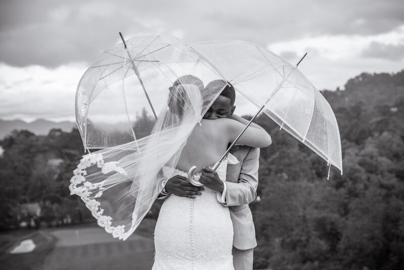 Bride in white dress holding transparent umbrellas with veil flowing in wind, photographed outdoors in black and white.