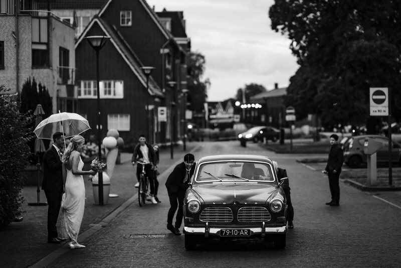 Black and white photograph of a vintage car on a wet street with pedestrians and period buildings in the background.