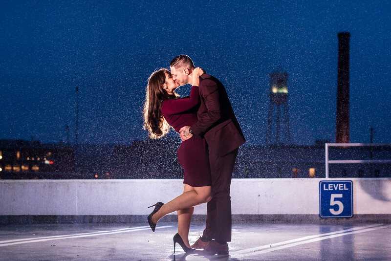 Couple kissing on a rooftop parking level at night, with rain falling and city lights in the background.