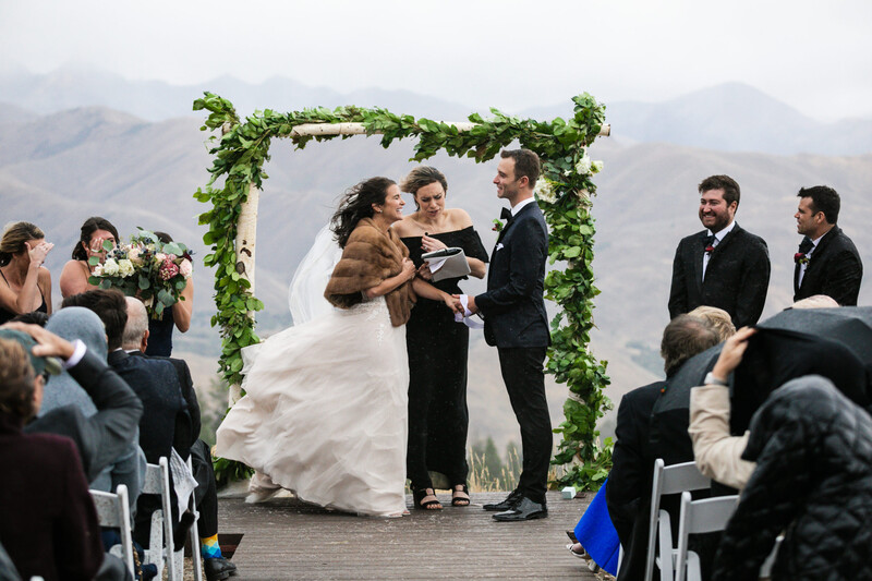 Bride and groom exchange vows at outdoor ceremony beneath greenery-decorated arch with mountainous backdrop.