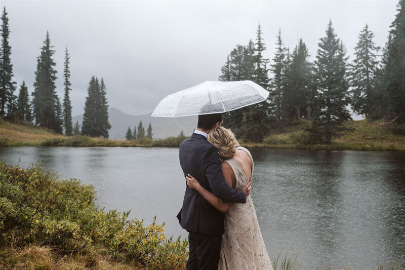 Couple embracing under a white umbrella beside a calm river surrounded by evergreen forest and overcast sky.