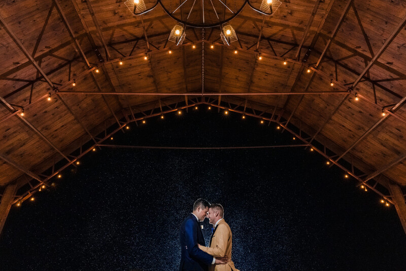 Couple kissing under wooden barn ceiling with warm overhead lighting and dark night sky backdrop.