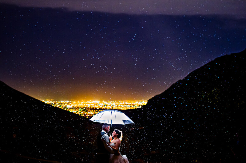 Couple under illuminated umbrella in rain with starry sky and city lights framed by dark mountains.