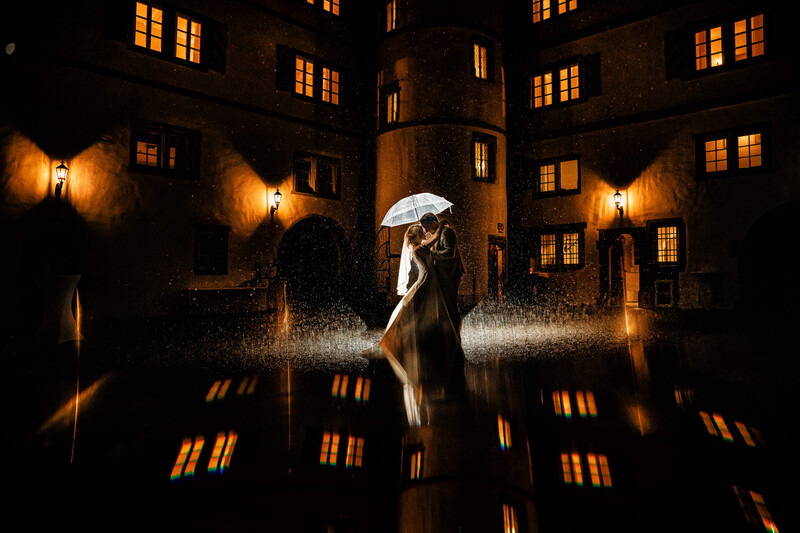 Person holding white umbrella in heavy rain at night in courtyard surrounded by illuminated building windows.