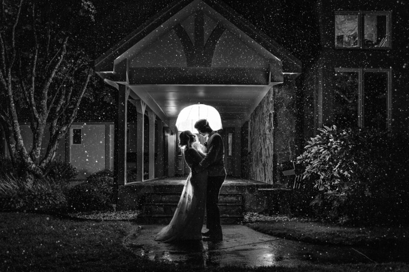 Couple kissing under an overhead light fixture on a covered porch during rain at night.