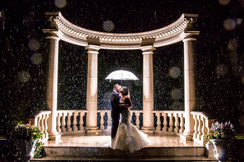 Bride and groom dancing under a white umbrella in heavy rain beneath a classical stone gazebo with columns.