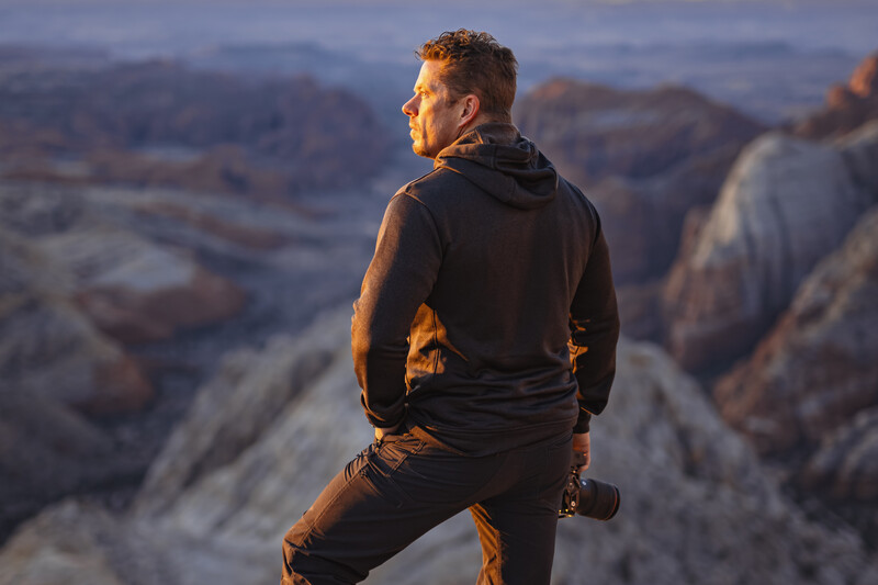 Man in black hoodie perched on rocky outcrop overlooking expansive canyon landscape at golden hour.