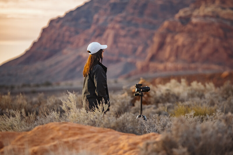 Woman in white cap and dark jacket standing in desert scrubland with tripod-mounted camera in foreground, layered mountains in background at golden hour.