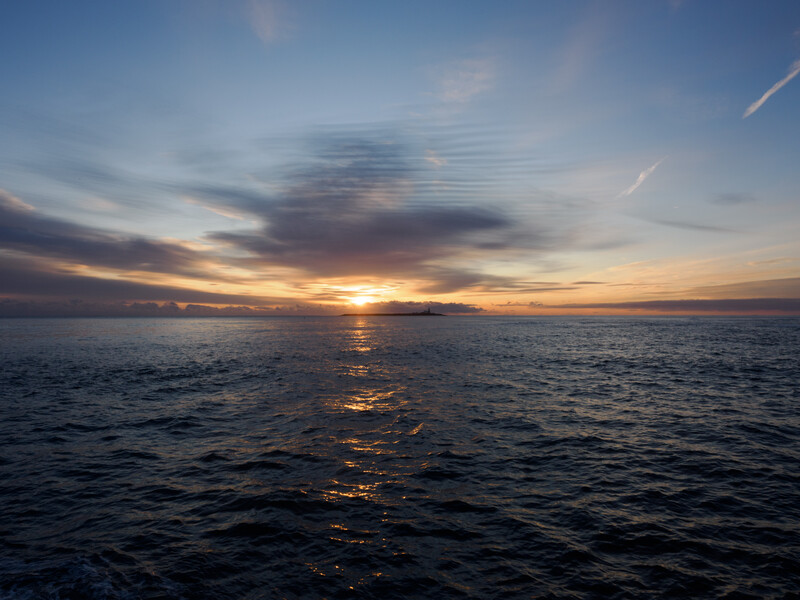 Golden sunset over calm ocean water with distant landmass silhouetted against colorful sky.