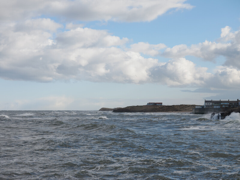 Coastal landscape with rough ocean waves, rocky outcrop with structures, and cloudy sky.