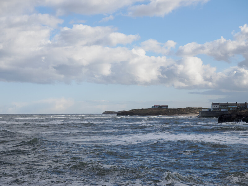 Coastal seascape with rolling waves, buildings on a distant headland, and partly cloudy sky.