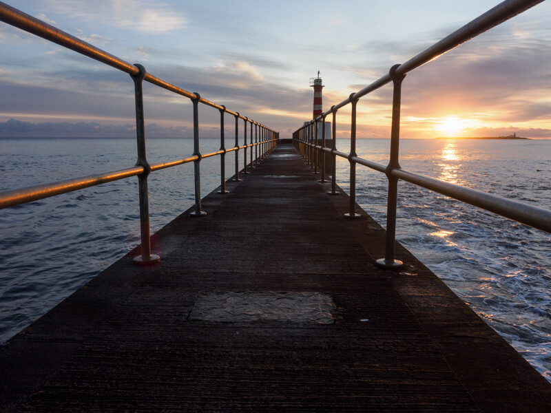 Long wooden pier with metal railings extending toward a lighthouse at sunset over calm water.