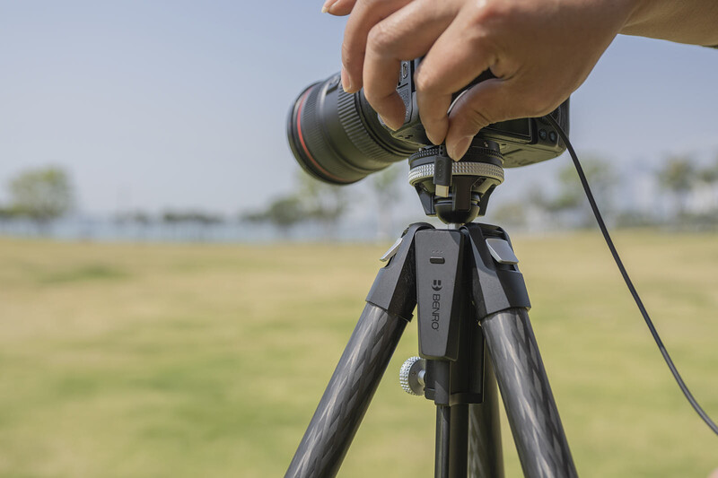 Hands mounting a telephoto lens onto a camera on a tripod in an open field.
