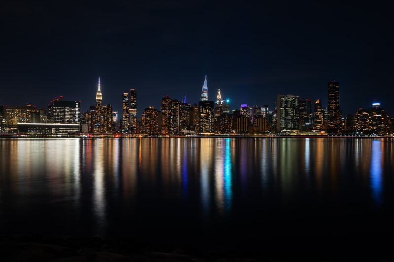 Manhattan skyline at night with illuminated skyscrapers reflected in dark water.