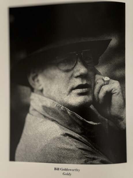 Black and white portrait of a man wearing glasses and a hat, hand raised to his face in a thoughtful pose.