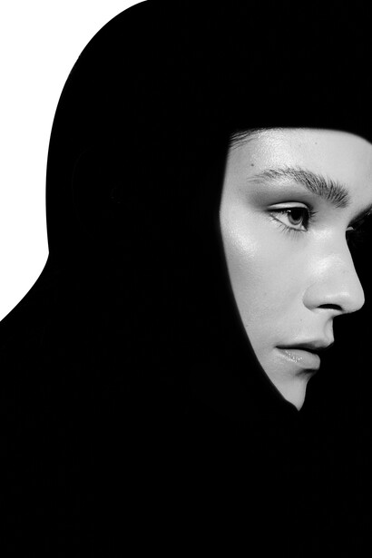 Profile portrait of a person wearing a black hood against a dark background.