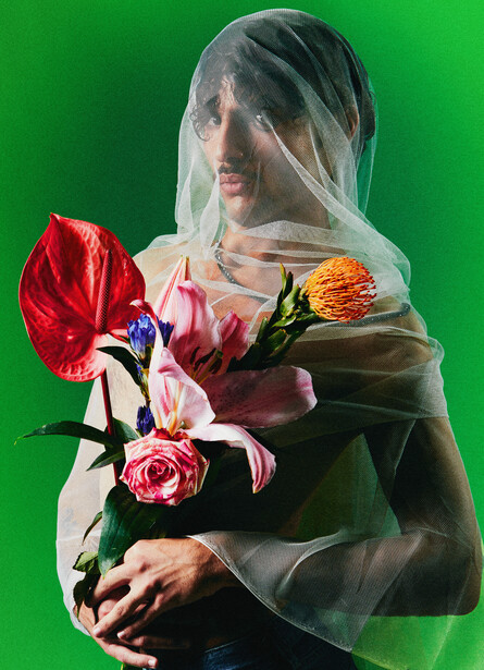 Person wearing sheer veil holding vibrant bouquet of red, pink, and yellow flowers against green background.