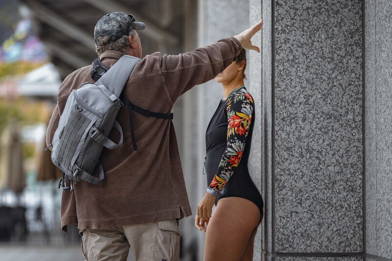 Photographer with gray backpack directing a model wearing colorful sleeve tattoo against concrete wall.