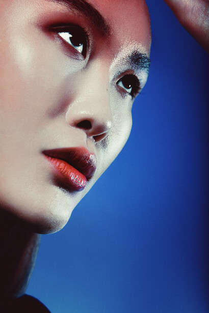Profile of a woman's face with bold burgundy eyeshadow and coral-red lips against a deep blue background.