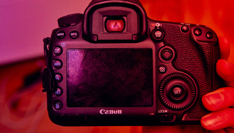 Canon DSLR camera viewed from the rear, illuminated by red and magenta gel lighting against a colored background.