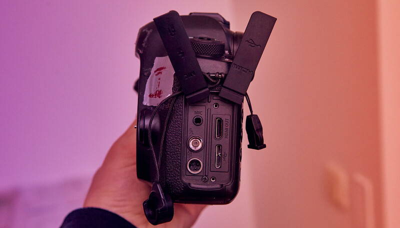 Handheld video camera held up to camera, showing rear panel with buttons and ports against pink and purple gradient lighting.