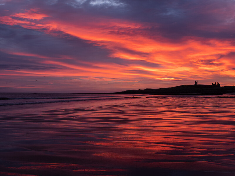 Dramatic sunset over a calm ocean with silhouetted landmass and structures on the horizon.