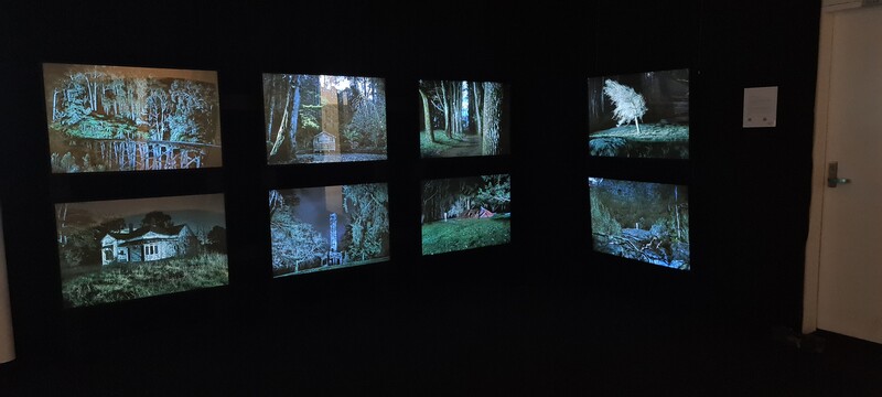 Gallery wall display of eight illuminated video monitors showing nature scenes with water, trees, and architectural elements against black walls.