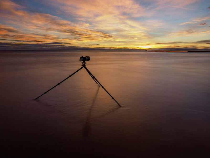 Tripod standing in shallow water at sunset with golden sky and calm sea.