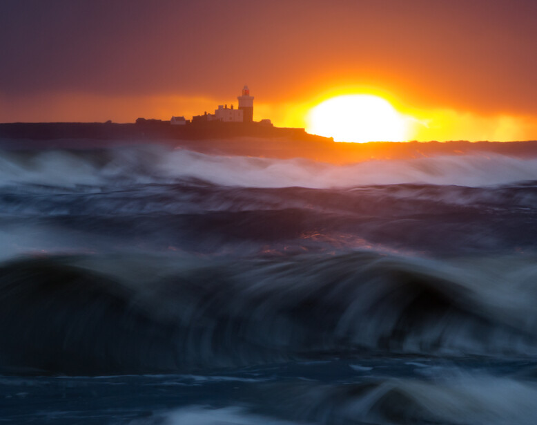 Dramatic coastal sunrise with intense golden sun, rolling waves with motion blur, and silhouetted buildings on the shoreline.