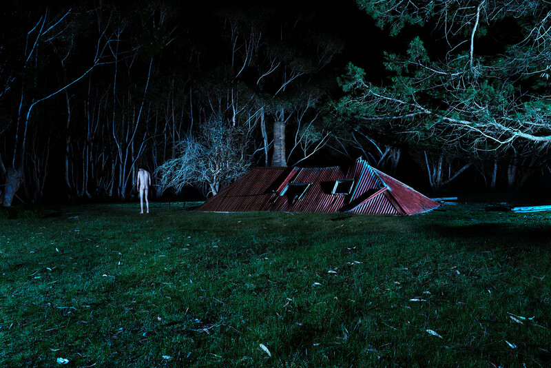 Person standing in a dark field facing an illuminated abandoned structure surrounded by bare trees.
