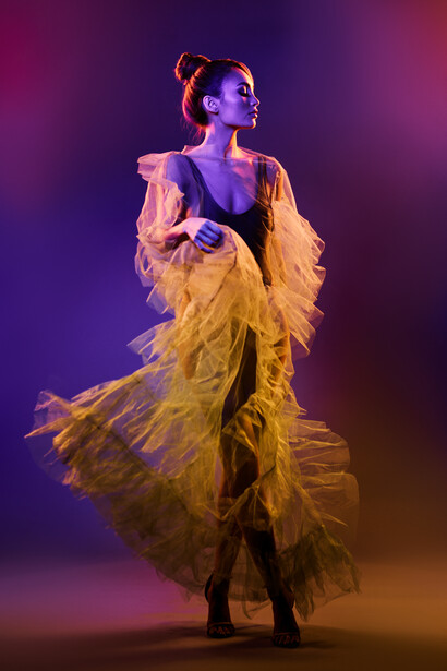 Woman in flowing yellow garment posing against purple and amber gradient backdrop with dramatic lighting.