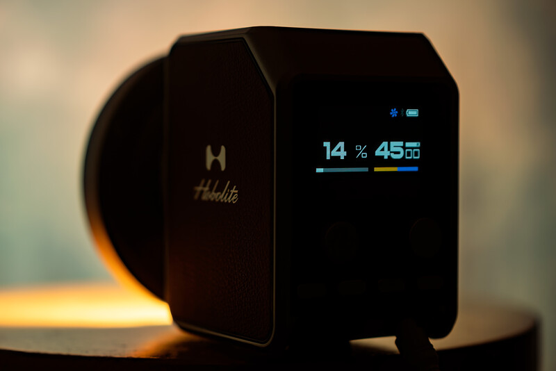 Compact black device with illuminated display showing battery percentage and time against warm studio lighting.