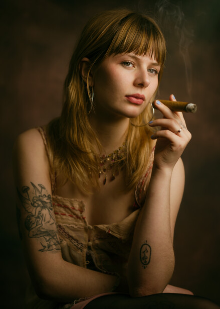 Woman with tattoos holding a cigarette, wearing layered clothing against a dark background.
