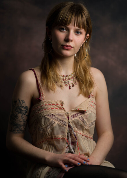 Woman with long blonde hair and bangs wearing an ornate beaded necklace and patterned sleeveless top.