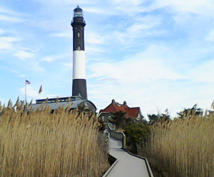 Striped lighthouse with keeper's house viewed through tall marsh grasses along a wooden boardwalk.