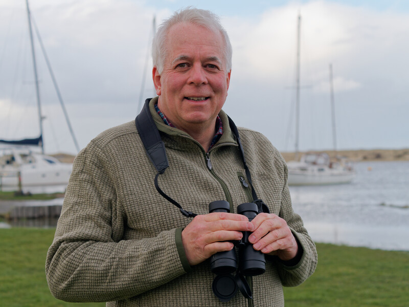 Older man holding binoculars at a waterfront with sailboats in the background.
