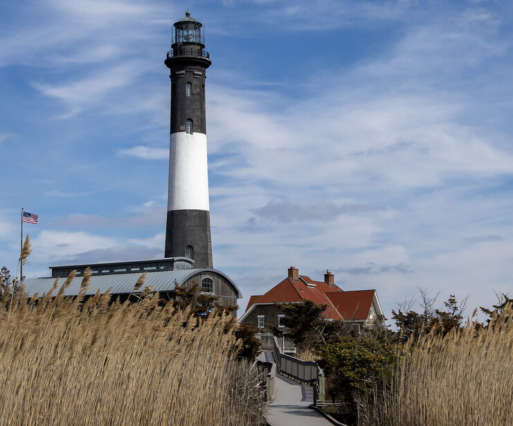 Historic lighthouse with black and white striped tower stands behind coastal buildings surrounded by golden marsh grasses.