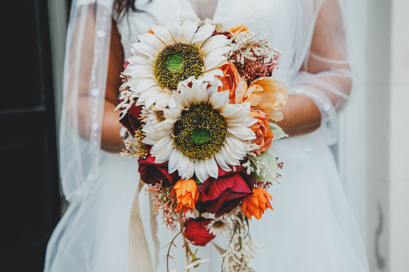 Bride in white dress holding cascading bouquet of white daisies, orange and red flowers with dried foliage.