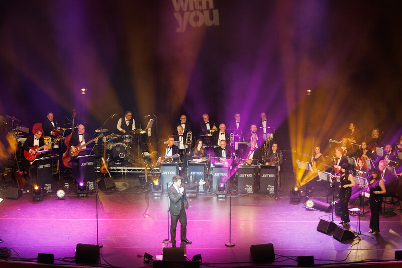 Large orchestra and band performing on stage with dynamic purple and gold lighting design.