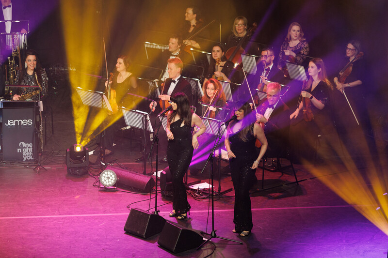 Orchestra performing on stage with dramatic purple and golden uplighting during a concert.