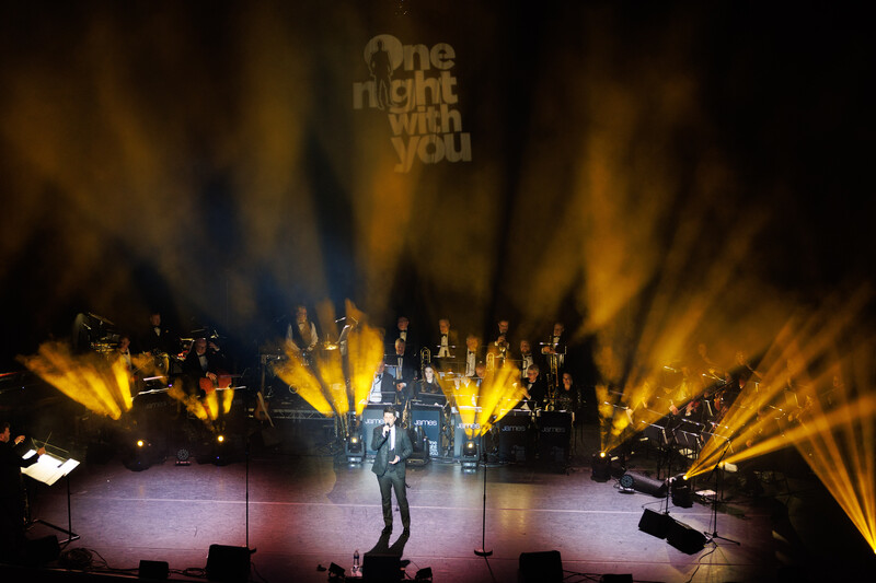 Solo performer on stage with dramatic golden and blue spotlights radiating outward during a concert.