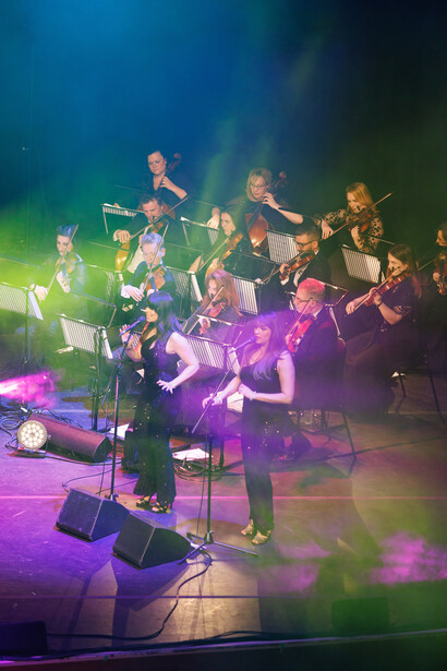 Orchestra ensemble performing on stage with vibrant purple, green, and blue stage lighting.
